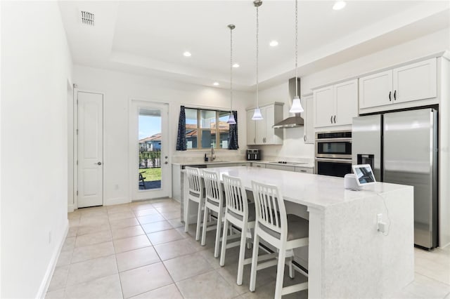 kitchen featuring visible vents, stainless steel appliances, a raised ceiling, wall chimney exhaust hood, and a center island