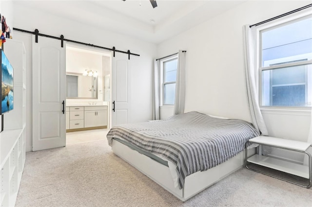 bedroom with ensuite bath, a barn door, a raised ceiling, light colored carpet, and ceiling fan