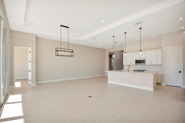 kitchen featuring baseboards, a tray ceiling, a kitchen island with sink, white cabinets, and stainless steel microwave