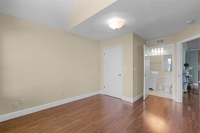 unfurnished bedroom featuring visible vents, ensuite bathroom, a textured ceiling, wood finished floors, and baseboards