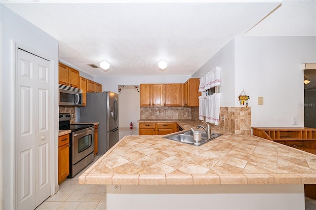 kitchen with brown cabinets, a sink, tile countertops, appliances with stainless steel finishes, and a peninsula