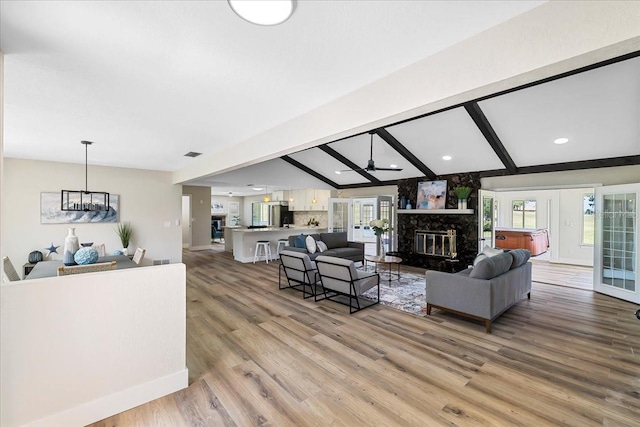 living area featuring visible vents, ceiling fan with notable chandelier, a stone fireplace, light wood finished floors, and vaulted ceiling with beams