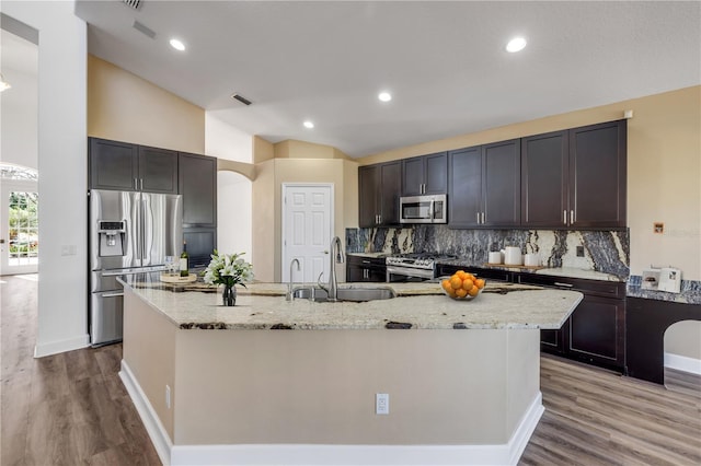 kitchen with light stone countertops, a kitchen island with sink, a sink, stainless steel appliances, and vaulted ceiling