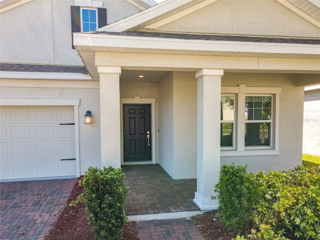 view of exterior entry with stucco siding, decorative driveway, and roof with shingles