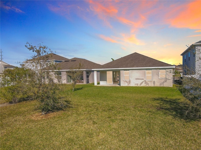 back of property at dusk featuring stucco siding, a lawn, and roof with shingles