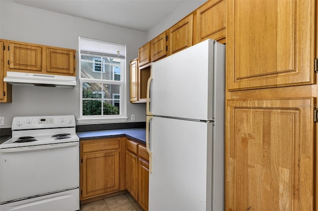 kitchen with dark countertops, under cabinet range hood, light tile patterned floors, brown cabinetry, and white appliances