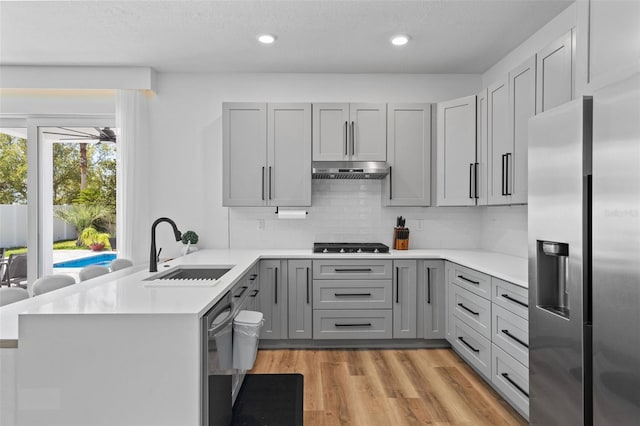 kitchen featuring gray cabinets, a sink, under cabinet range hood, appliances with stainless steel finishes, and a peninsula