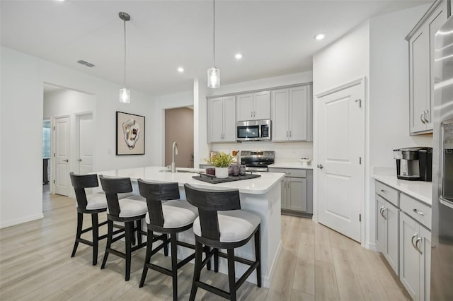 kitchen with a kitchen island with sink, gray cabinets, appliances with stainless steel finishes, a kitchen breakfast bar, and light wood-type flooring