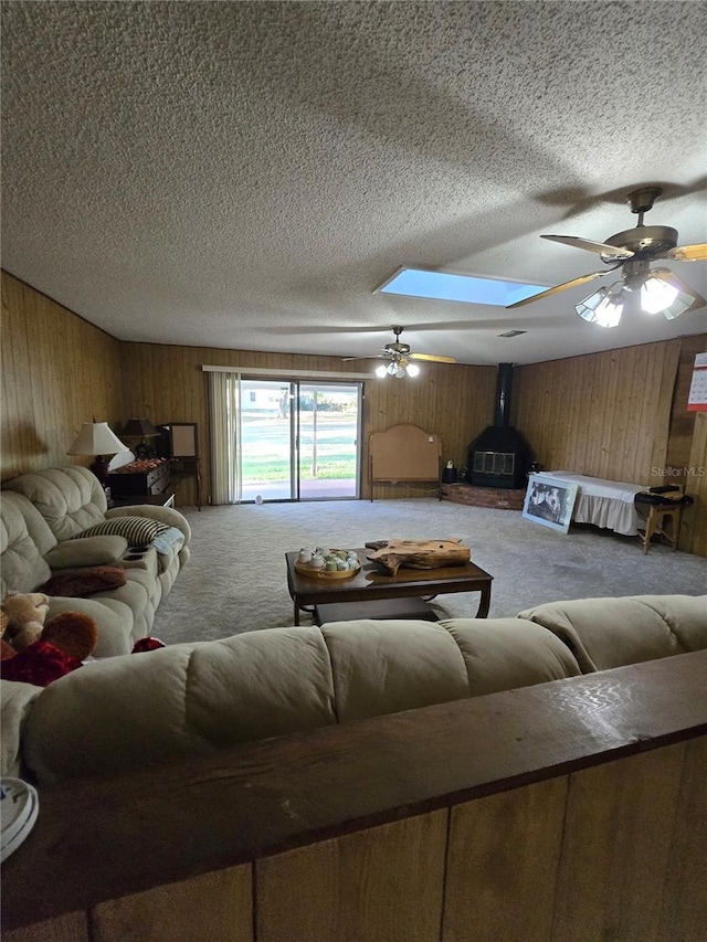 living room featuring wooden walls, carpet flooring, a skylight, a wood stove, and a ceiling fan