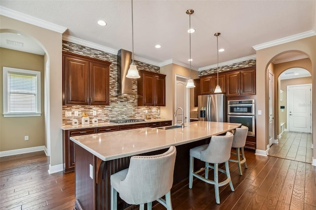 kitchen with wall chimney range hood, decorative backsplash, appliances with stainless steel finishes, arched walkways, and a sink