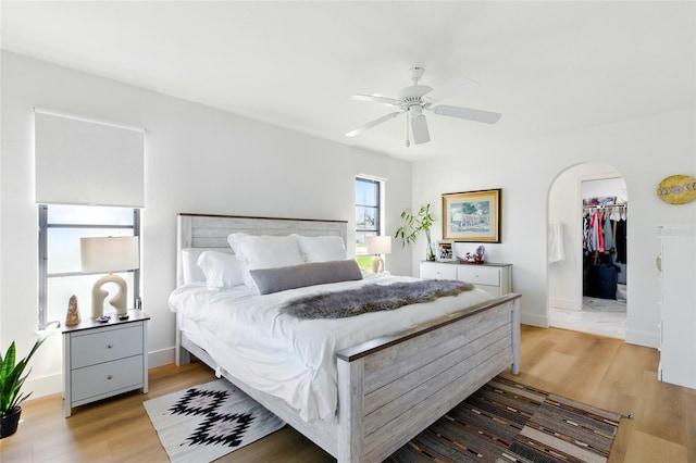 bedroom featuring a walk in closet, light wood-style flooring, arched walkways, baseboards, and ceiling fan