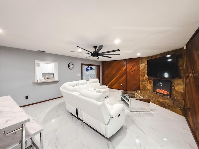 living area with marble finish floor, wooden walls, a stone fireplace, baseboards, and ceiling fan