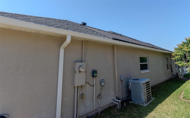 view of side of home with stucco siding, a lawn, central AC, roof with shingles, and solar panels