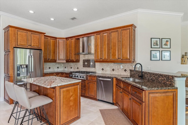 kitchen with a sink, appliances with stainless steel finishes, brown cabinetry, and wall chimney range hood