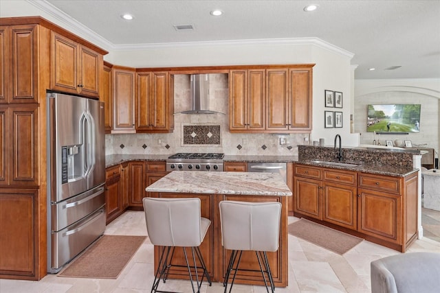 kitchen featuring a peninsula, a sink, stainless steel appliances, wall chimney exhaust hood, and brown cabinets