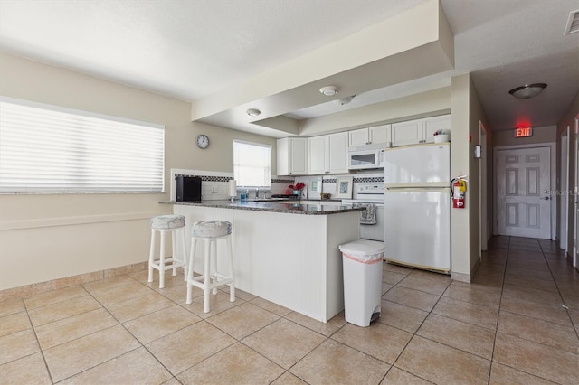 kitchen featuring a kitchen bar, white appliances, a peninsula, light tile patterned flooring, and white cabinets