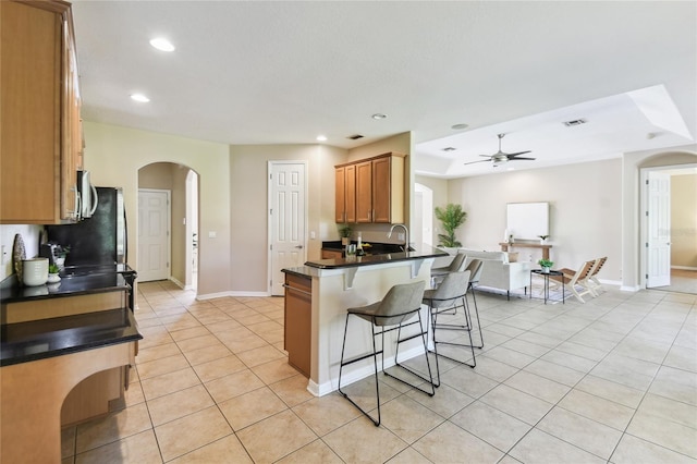 kitchen with a breakfast bar area, light tile patterned floors, dark countertops, arched walkways, and ceiling fan
