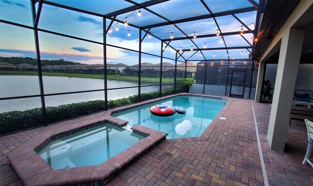 pool at dusk featuring glass enclosure, a water view, a pool with connected hot tub, and a patio area