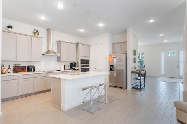 kitchen featuring light wood-style flooring, a breakfast bar, stainless steel appliances, wall chimney exhaust hood, and a center island with sink