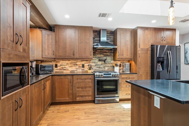 kitchen featuring wall chimney range hood, appliances with stainless steel finishes, dark countertops, light wood-type flooring, and decorative backsplash