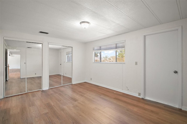 unfurnished bedroom featuring two closets, baseboards, a textured ceiling, visible vents, and wood finished floors