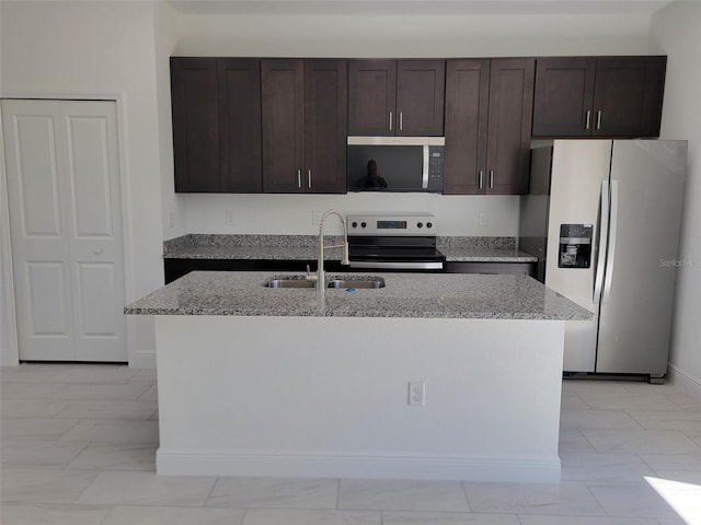 kitchen featuring stainless steel appliances, a sink, an island with sink, light stone counters, and dark brown cabinets