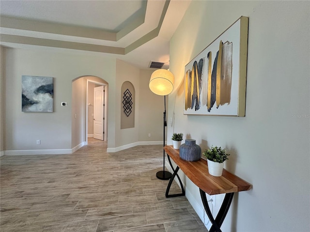 hall with a tray ceiling, wood finished floors, visible vents, arched walkways, and baseboards