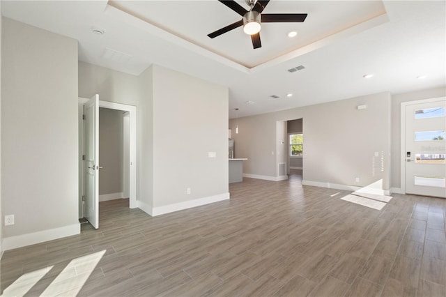 unfurnished living room with a tray ceiling, light wood-style flooring, baseboards, visible vents, and ceiling fan