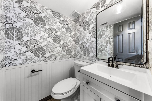 half bathroom with visible vents, wallpapered walls, toilet, vanity, and wainscoting