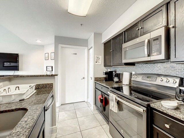kitchen with stainless steel appliances, dark brown cabinets, dark stone countertops, light tile patterned flooring, and a textured ceiling