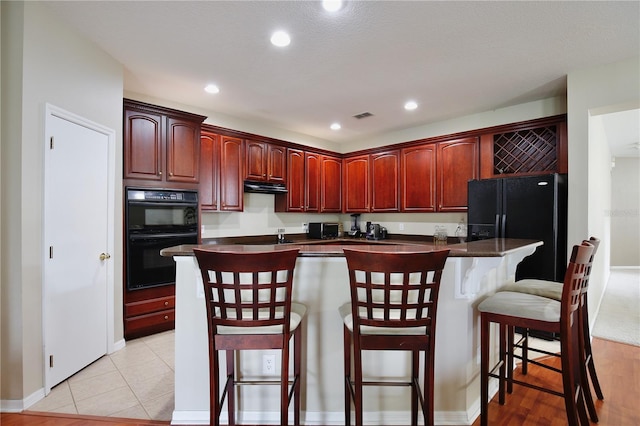 kitchen with under cabinet range hood, black appliances, visible vents, dark countertops, and reddish brown cabinets