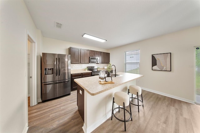 kitchen with a kitchen breakfast bar, dark brown cabinetry, a center island with sink, light wood-style flooring, and stainless steel appliances