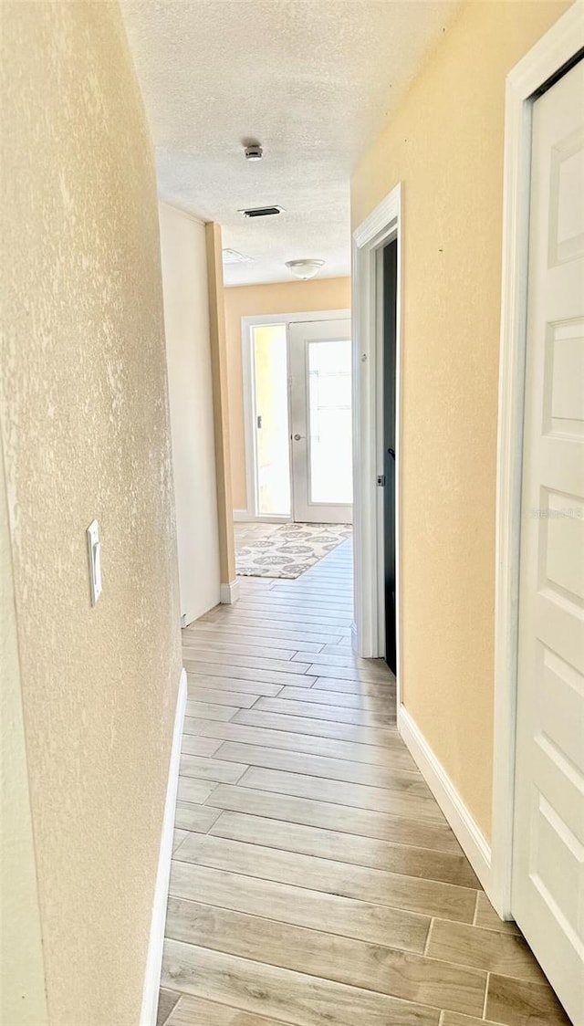 corridor featuring light wood-type flooring, a textured wall, and a textured ceiling
