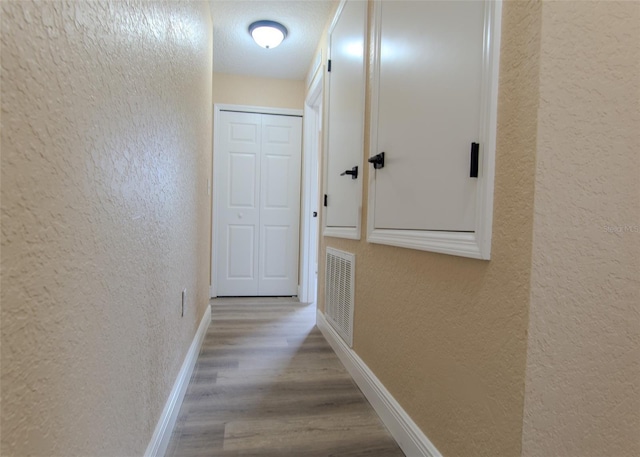 hallway featuring a textured wall, visible vents, baseboards, and wood finished floors