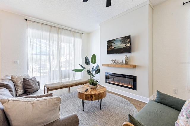 living room with a textured ceiling, a glass covered fireplace, baseboards, wood finished floors, and ceiling fan