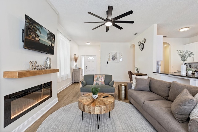 living area featuring arched walkways, a textured ceiling, a glass covered fireplace, a ceiling fan, and wood finished floors