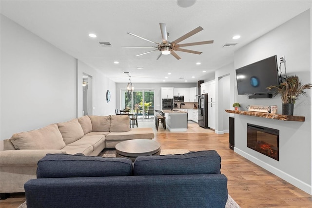 living area featuring recessed lighting, light wood-type flooring, ceiling fan with notable chandelier, baseboards, and a glass covered fireplace