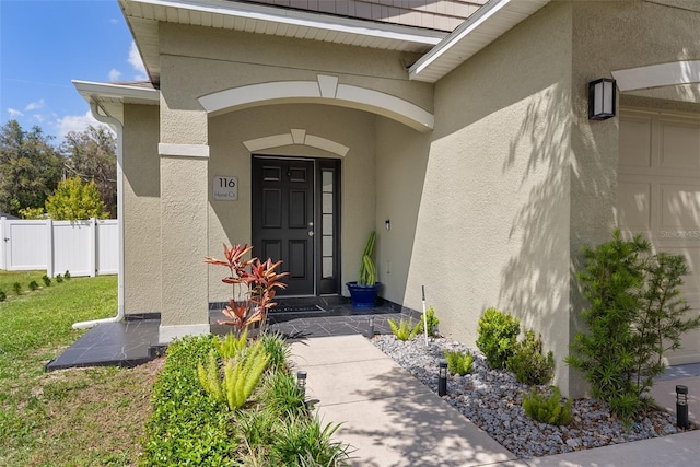 view of exterior entry with a garage, stucco siding, and fence
