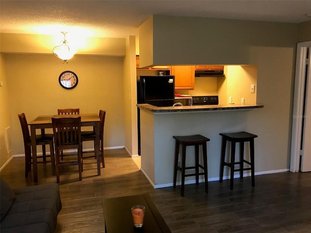 kitchen featuring range, freestanding refrigerator, dark countertops, under cabinet range hood, and a breakfast bar area