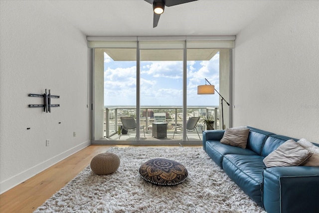 living area featuring plenty of natural light, ceiling fan, wood finished floors, a wall of windows, and baseboards