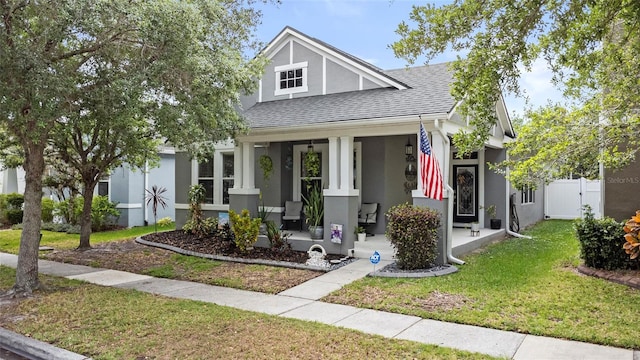 bungalow featuring a front lawn, covered porch, fence, stucco siding, and roof with shingles