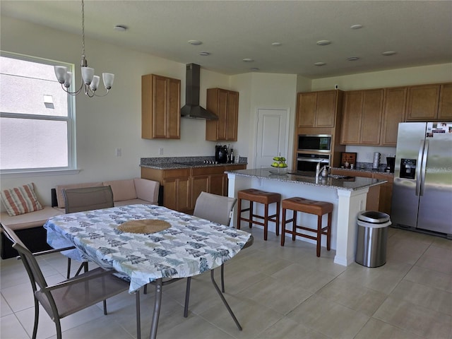 1764 Stamen Dr, Kissimmee, FL 34744 - kitchen featuring an island with sink, brown cabinetry, wall chimney range hood, appliances with stainless steel finishes, and a notable chandelier