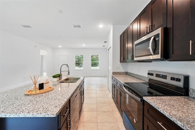 kitchen with stainless steel appliances, a sink, light stone counters, dark brown cabinets, and a center island with sink