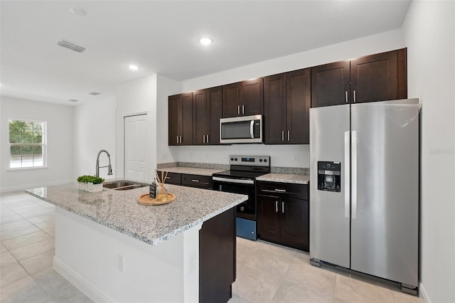kitchen featuring appliances with stainless steel finishes, a sink, a center island with sink, dark brown cabinetry, and light stone countertops