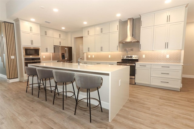 kitchen with a large island with sink, wall chimney range hood, appliances with stainless steel finishes, and light wood-style floors