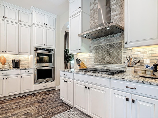 kitchen featuring white cabinets, arched walkways, wall chimney range hood, stainless steel appliances, and backsplash
