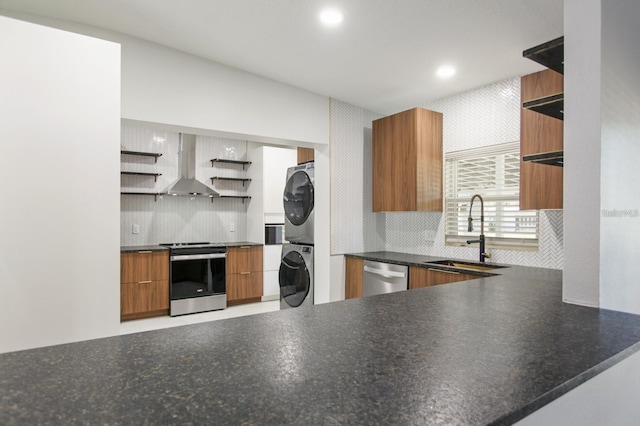 kitchen with open shelves, a sink, wall chimney range hood, appliances with stainless steel finishes, and stacked washer / dryer