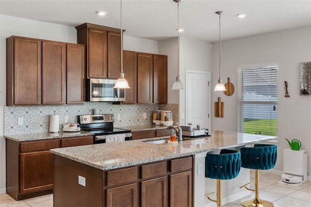 kitchen with light tile patterned floors, appliances with stainless steel finishes, an island with sink, light stone countertops, and backsplash