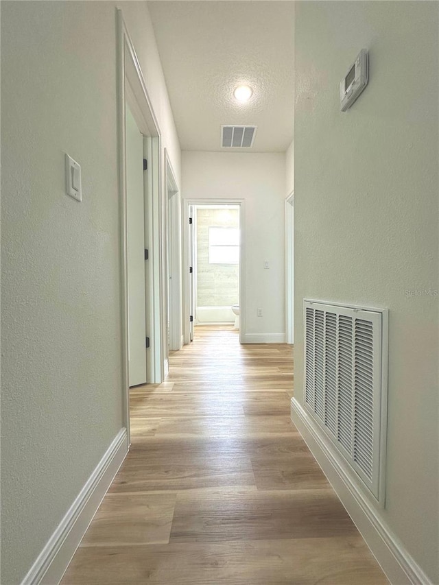 hallway featuring baseboards, a textured wall, light wood-style flooring, and visible vents