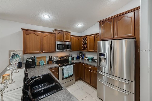 kitchen with stainless steel appliances, light tile patterned floors, brown cabinetry, vaulted ceiling, and a textured ceiling